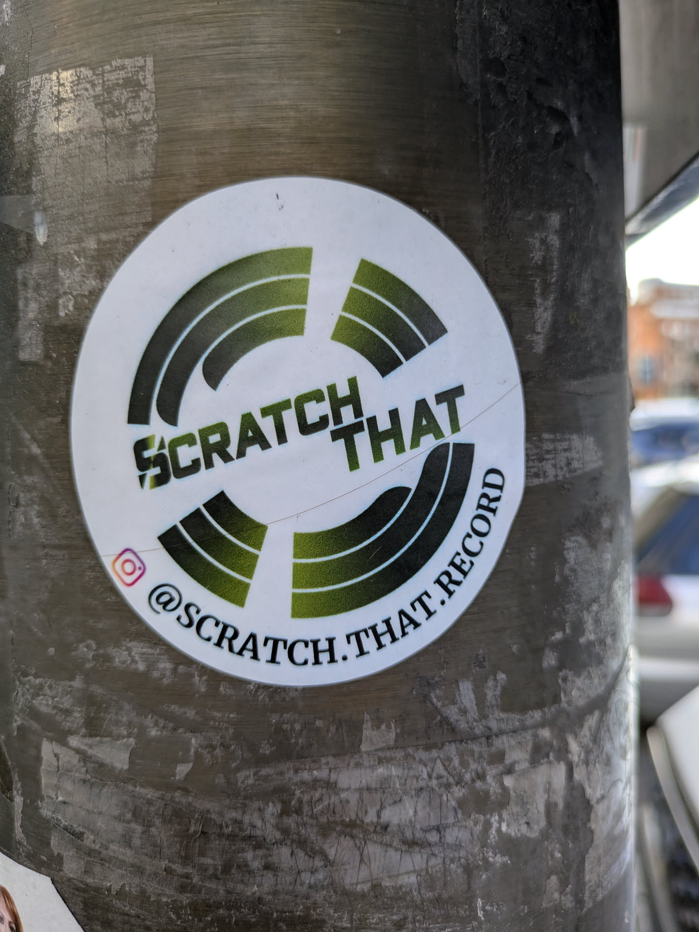 This is a white, round sticker with the words 'Scratch That' in bold black letters stacked on top of 'Record'. A green gradient symbol resembling rotating records surrounds the text. An Instagram icon is present with the username @scratch.that.record.