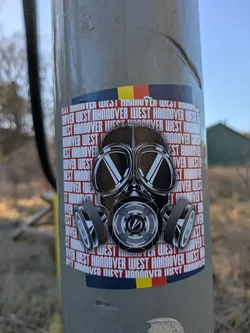 This sticker features a black and white drawing of a gas mask on a background with repeating text "West Hannover". The sticker also includes a stylized band of colors (black, red and yellow) at the top and bottom.