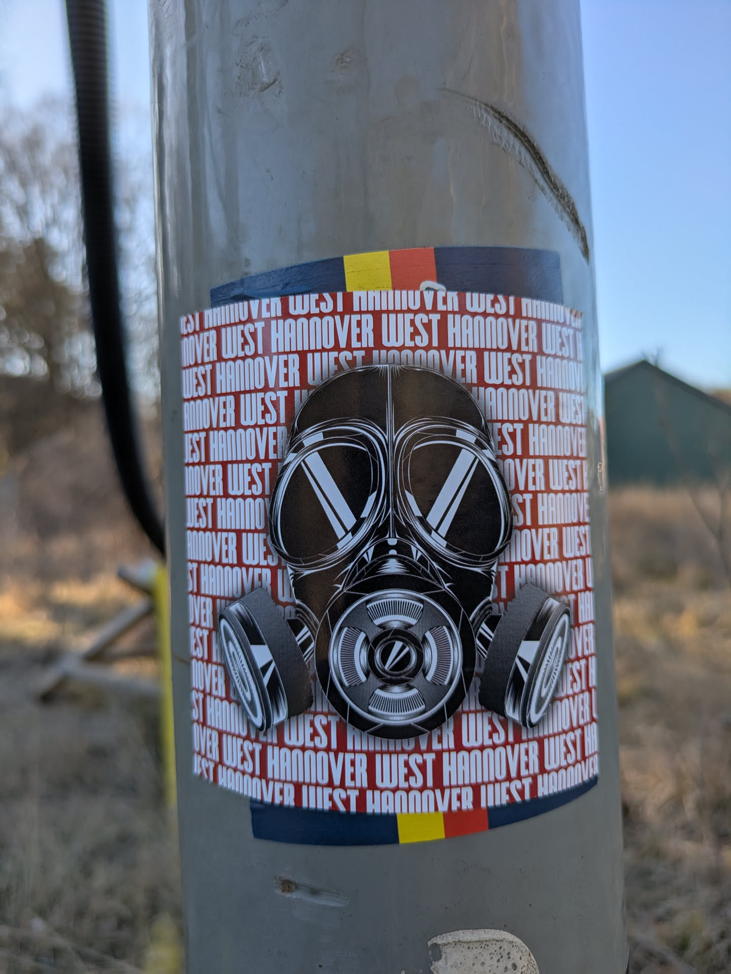 This sticker features a black and white drawing of a gas mask on a background with repeating text "West Hannover". The sticker also includes a stylized band of colors (black, red and yellow) at the top and bottom.