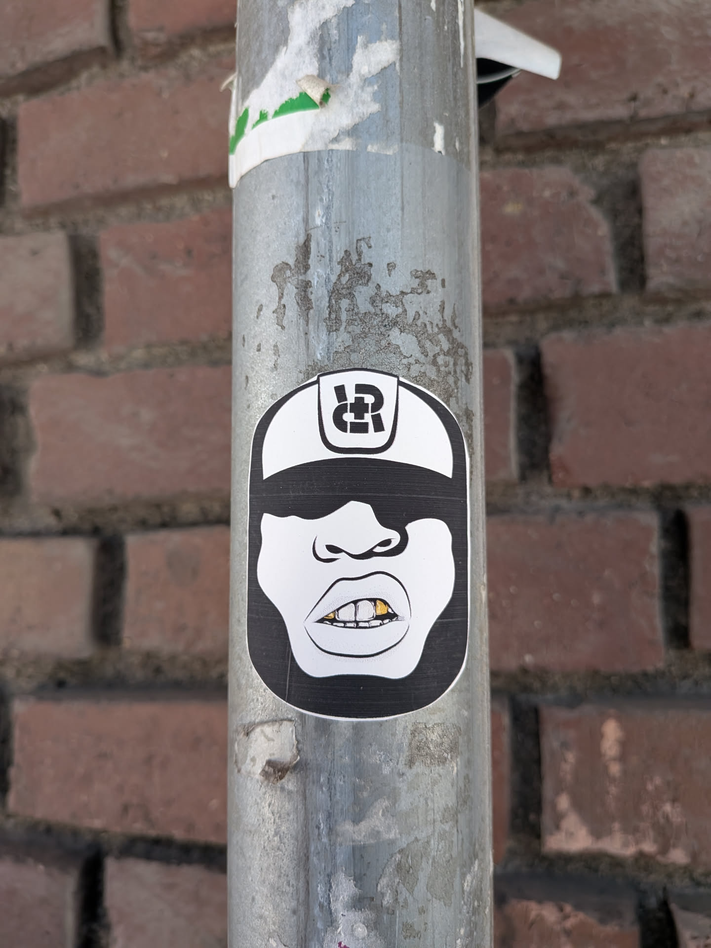 A monochrome, oval-shaped sticker featuring a stylized cartoon face. The character wears a white baseball cap with a black brim and a black, intertwined 'RP' or 'RD' logo on the front. Large black sunglasses obscure the eyes, and the face has prominent lips. The mouth is open, revealing white teeth, with two distinct gold teeth (grillz) on the bottom row. The overall design is bold and graphic, primarily in black and white with a touch of gold.