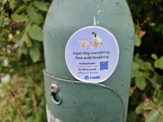 A round sticker featuring a woman picking up after her dog, promoting Lassie insurance with a discount code. The sticker is attached to a green pole outdoors.