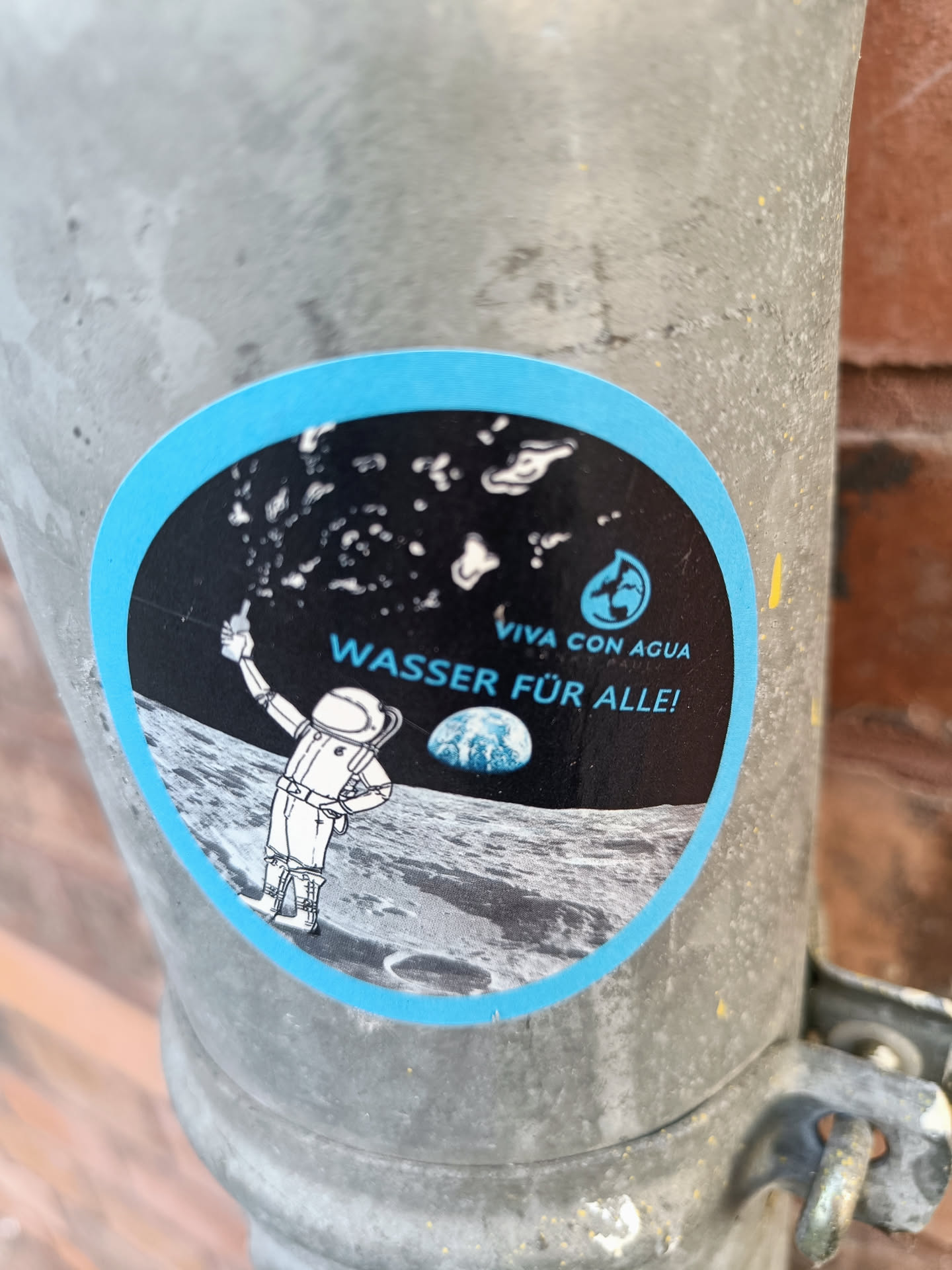 A round sticker featuring an illustration of an astronaut on the moon holding up water droplets. The text on the sticker says "VIVA CON AGUA" and "WASSER FÜR ALLE!" which translates to "Water for all!" in German. The sticker has a light blue border and a black background with a small globe image present. 