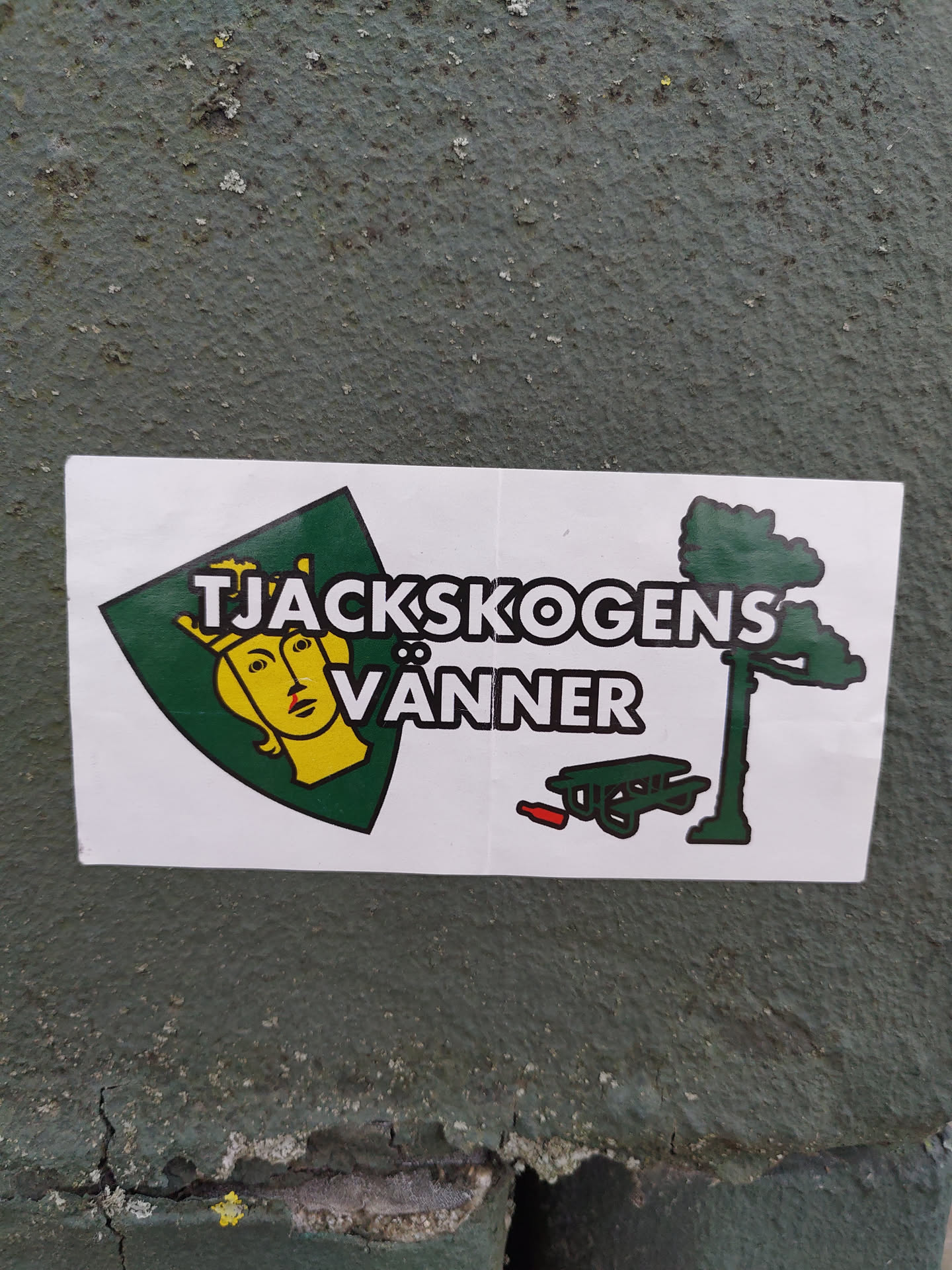 A sticker featuring a crest with a crowned woman's face, a pine tree, and a picnic table. The text on the sticker says "Tjackskogens Vänner".