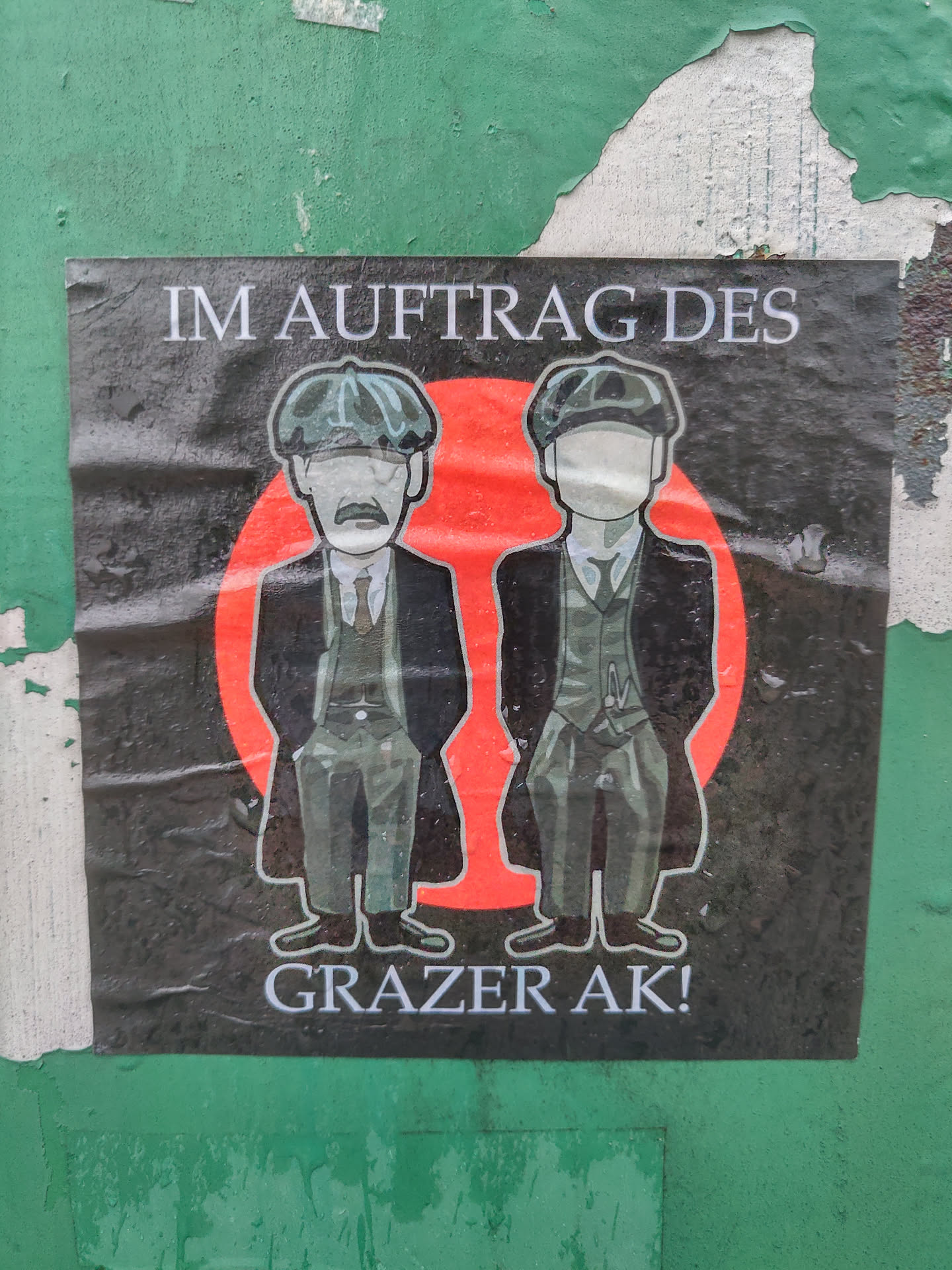 Sticker featuring two cartoon figures in suits and newsboy caps, set against a red circle.  Text at the top reads "Im Auftrag des" and at the bottom "Grazer AK!"