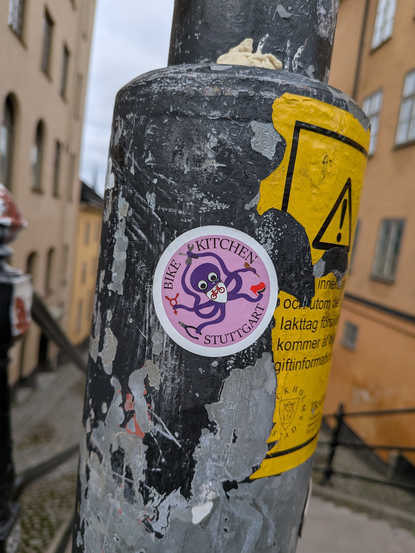 The sticker is circular with a pink border and a light pink background. In the center, a cartoonish purple octopus is depicted. The octopus has large, wide eyes and is wearing a white mask with a bicycle symbol on it. It has eight tentacles, two of which are holding tools: a wrench and a screwdriver. One tentacle is also holding a small red object. The words "BIKE KITCHEN" are written in a circular fashion above the octopus, and "STUTTGART" is written below it, both in a purple, slightly stylized font.