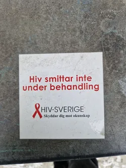 A small white sticker with red text and a red ribbon. The text on the sticker says "HIV does not spread during treatment" in Swedish. There is also a logo for HIV-Sverige, which says "Skyddar dig mot okunskap" (Protects you from ignorance).