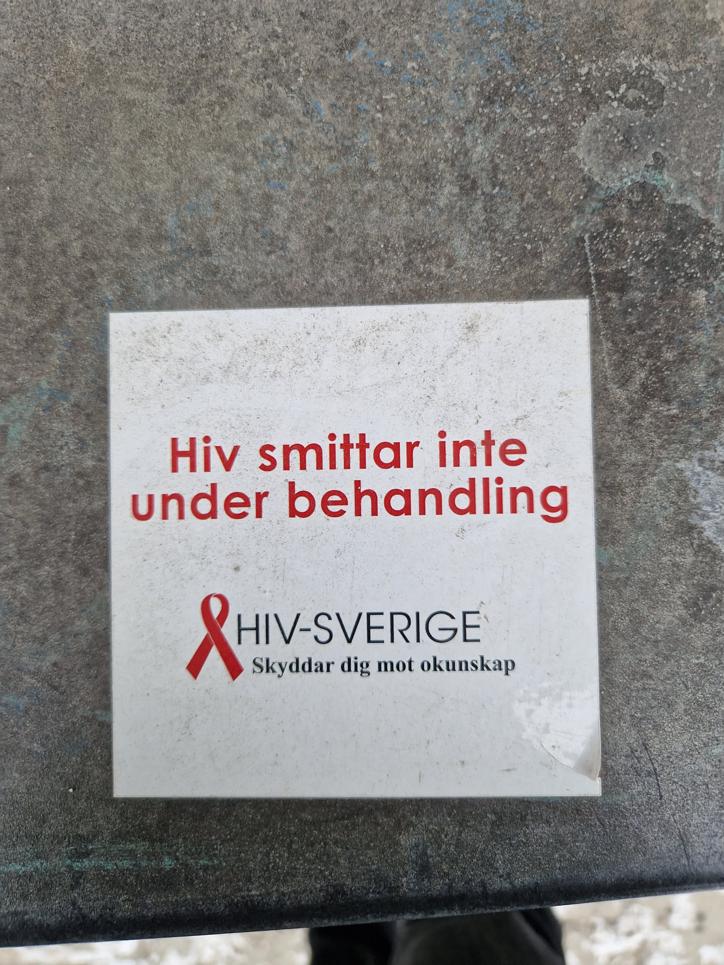 A small white sticker with red text and a red ribbon. The text on the sticker says "HIV does not spread during treatment" in Swedish. There is also a logo for HIV-Sverige, which says "Skyddar dig mot okunskap" (Protects you from ignorance).