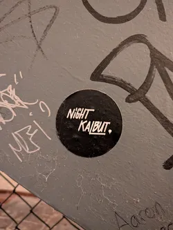 Street sticker A circular sticker affixed to a scratched and tagged grey metal or painted surface. The sticker features a solid black background with white stylized lettering reading 'NIGHT' above 'KALBUT.', followed by a small dot. The font has a dynamic, hand-drawn marker aesthetic.