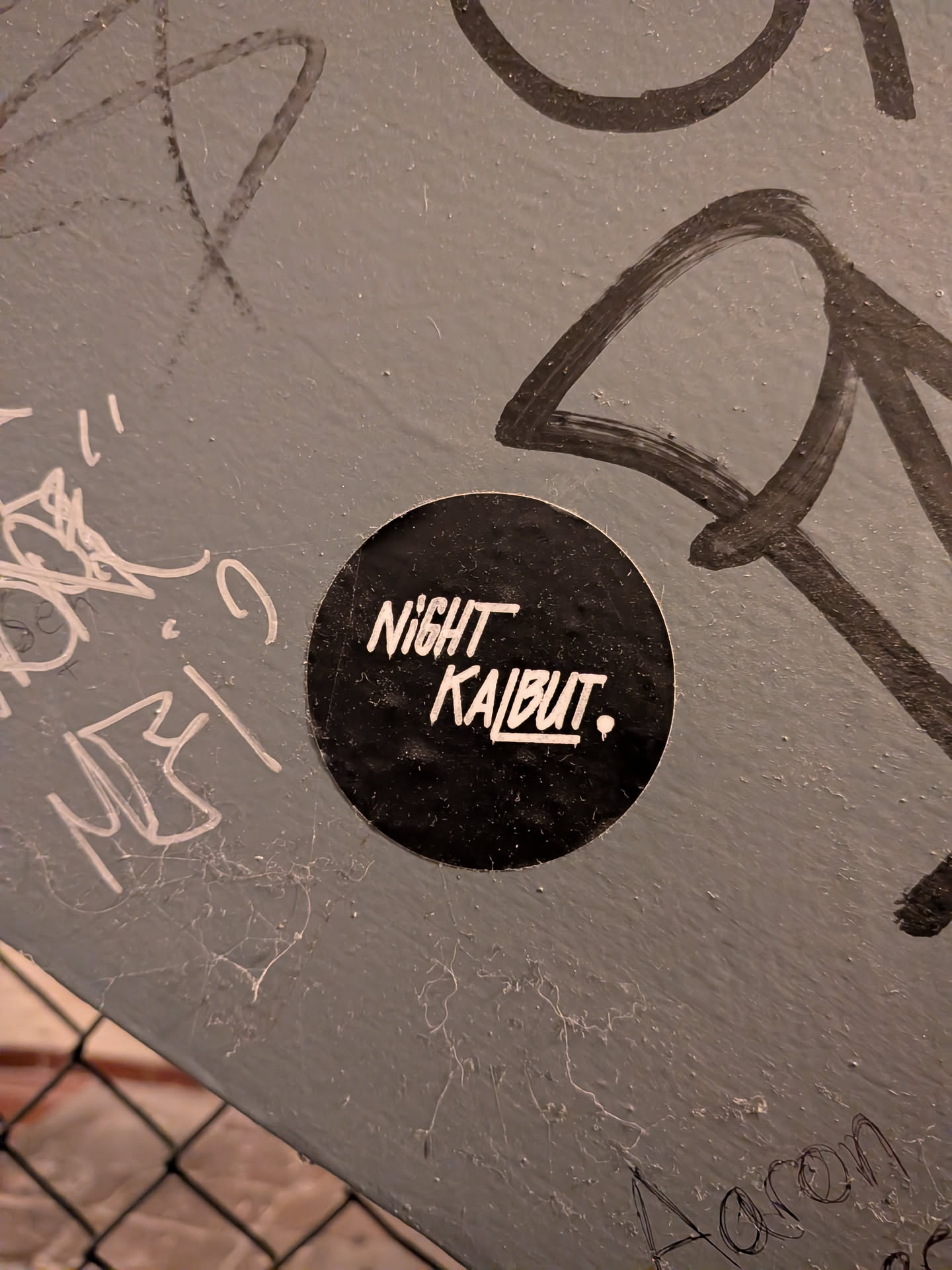 A circular sticker affixed to a scratched and tagged grey metal or painted surface. The sticker features a solid black background with white stylized lettering reading 'NIGHT' above 'KALBUT.', followed by a small dot. The font has a dynamic, hand-drawn marker aesthetic.
