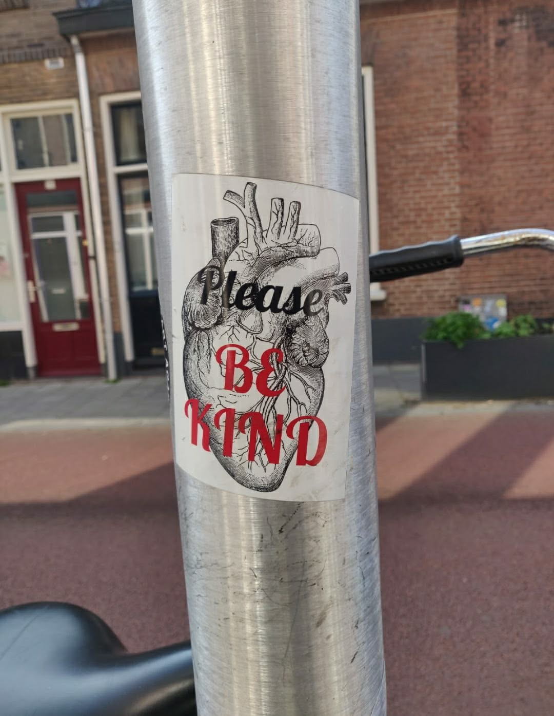 A sticker featuring an anatomical heart with the text 'Please Be Kind' in red lettering. The sticker is attached to a metal pole.