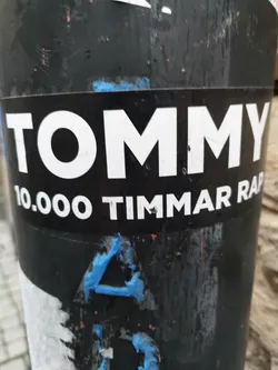 A black and white sticker with the text "TOMMY" in large white letters on top and "10.000 TIMMAR RAP" in smaller white letters underneath. The sticker is affixed to a dark-colored pole, which also has other stickers and markings on it. 