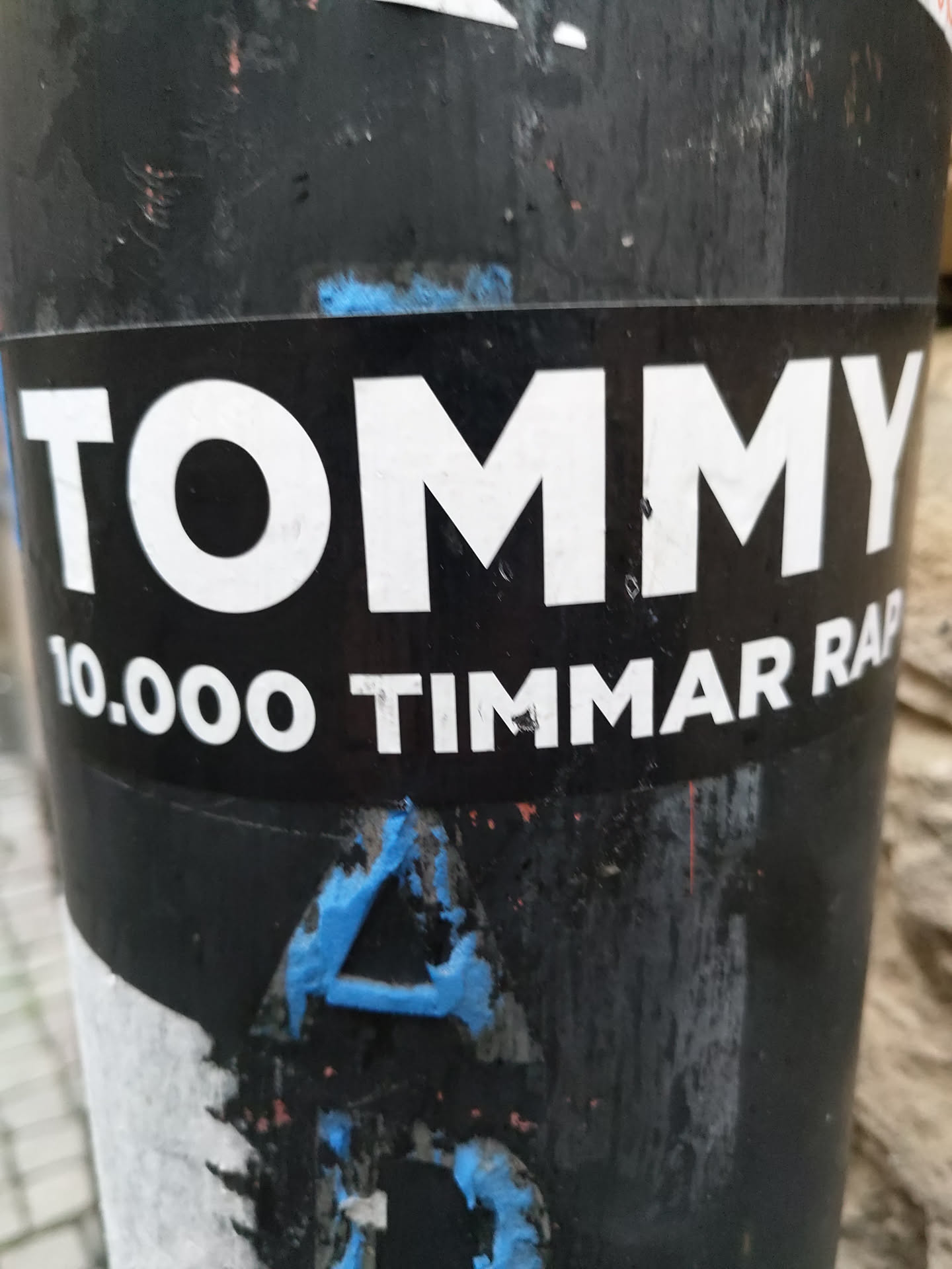 A black and white sticker with the text "TOMMY" in large white letters on top and "10.000 TIMMAR RAP" in smaller white letters underneath. The sticker is affixed to a dark-colored pole, which also has other stickers and markings on it. 
