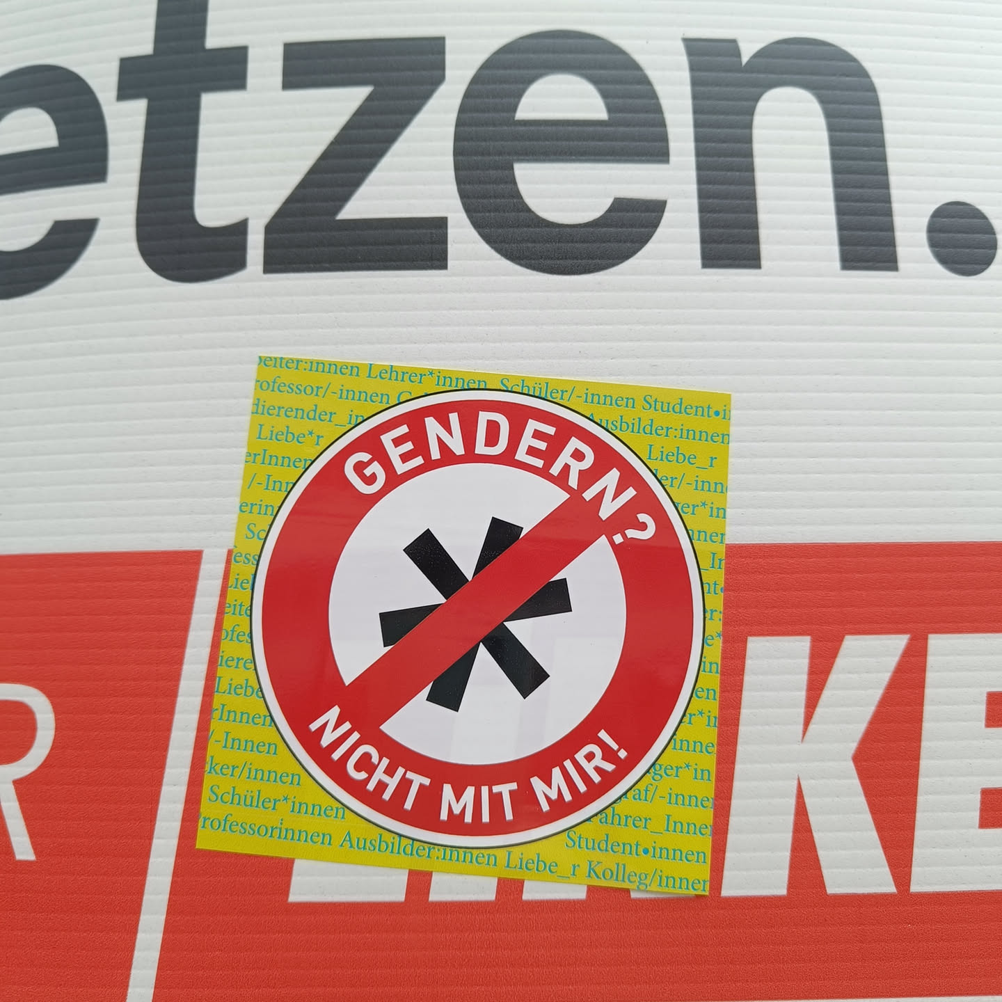 A round sticker with a red border and a white center.  A black asterisk with a red line across it is in the center. The text on the sticker says "GENDERN? NICHT MIT MIR!" which translates to "Gendering? Not with me!"
