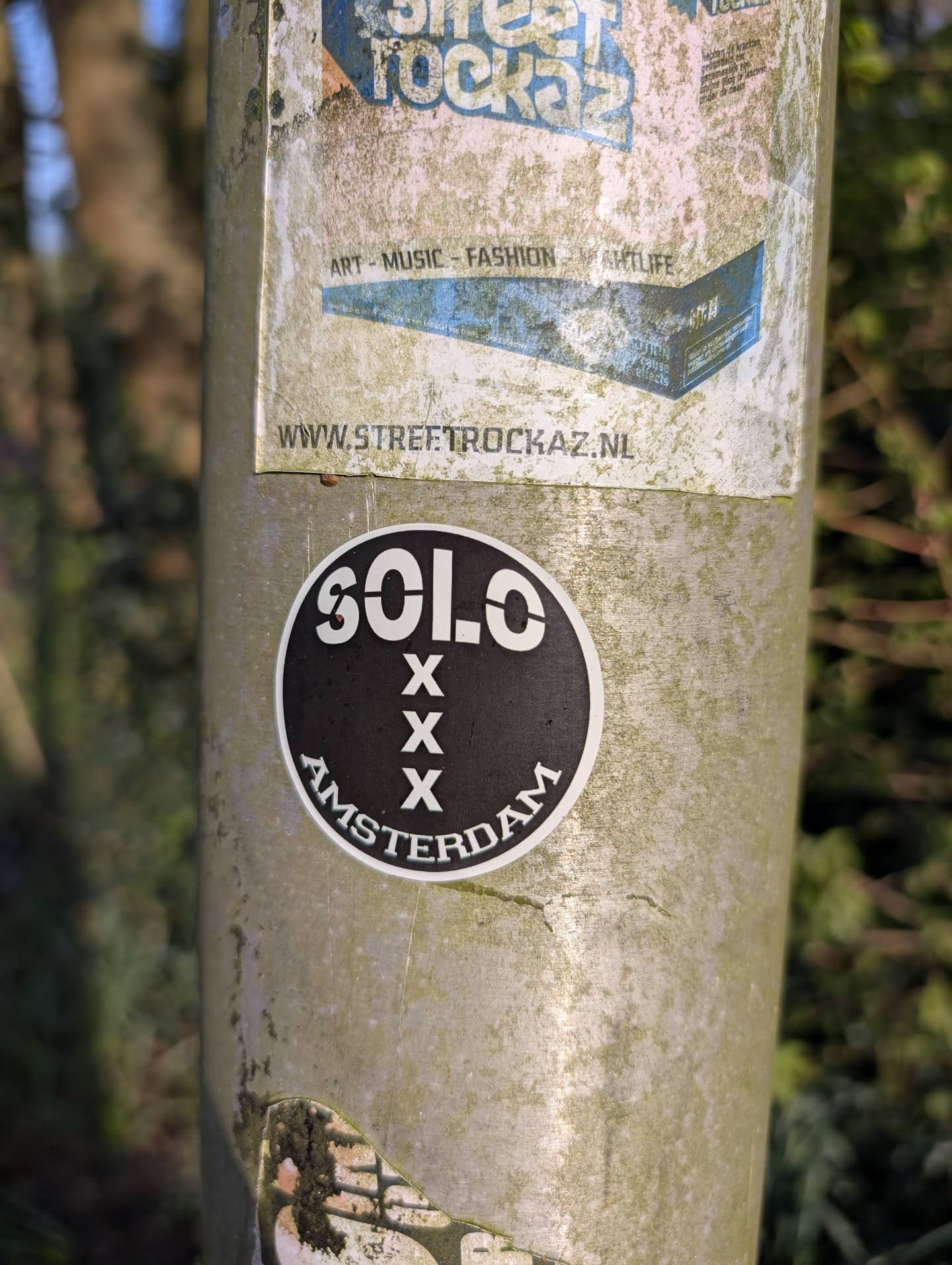 The sticker is round with a black background and white text and symbols. It reads "SOLO" at the top, followed by three "X" symbols stacked vertically, and "AMSTERDAM" at the bottom. The sticker is affixed to a weathered metal pole.