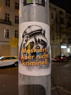 Street sticker A vertical sticker attached to a metal post features a black-and-white, woodcut-style illustration of a raccoon wearing a tweed flat cap and winking with one eye while tipping its cap. Overlaid on the lower half of the raccoon is bold yellow-orange text with dark drop shadows reading 'Maskiert. Aber nicht kriminell.'. In the bottom right corner, smaller green text identifies the creator as 'LAG Tierschutzpolitik Bündnis 90/Die Grünen Berlin'. The background of the sticker design is white.