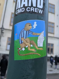 A sticker featuring a cartoon monkey wearing a blue and white striped shirt, sunglasses, smoking a cigarette, and reading a newspaper while sitting on the grass. 