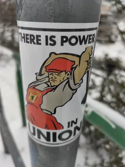 A sticker featuring a cartoon illustration of a person with a red hat and apron wielding a large hammer. The text on the sticker reads "There is power in union".  There is a small emblem on the apron.