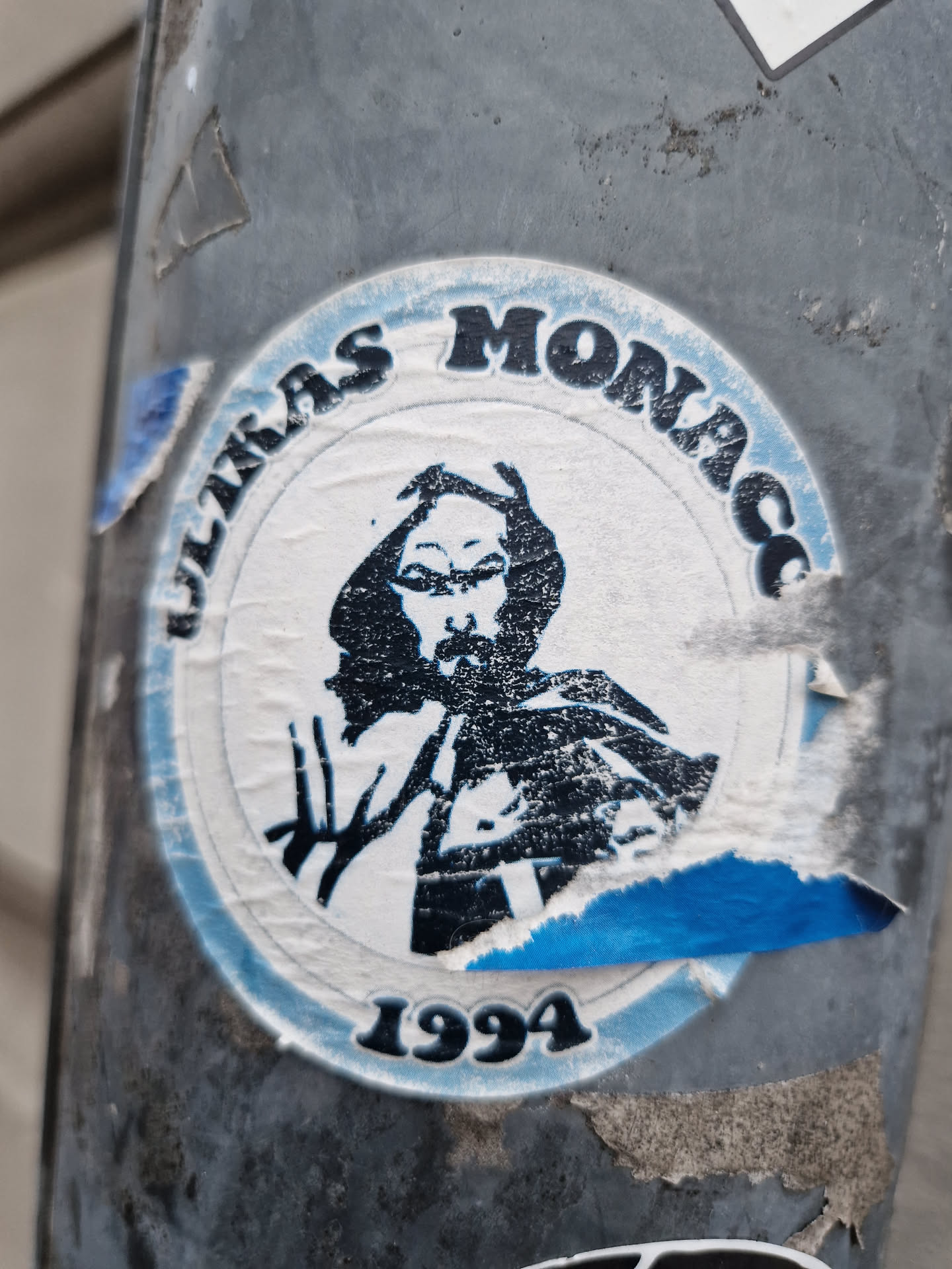 A worn sticker featuring a black and white illustration of a person, possibly a religious figure or historical figure, with the text "Ultras Monaco 1994" encircling the image. The sticker shows signs of wear and tear, with parts of it peeling off.