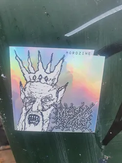 This sticker features a holographic design. The main image is a black and white drawing of a demonic or evil-looking figure wearing a crown.  There is some additional text at the bottom right, which appears to be a band logo.  The sticker is rectangular with a slight curve to the top right corner. The text 'MORDZINE.' is visible on the sticker.