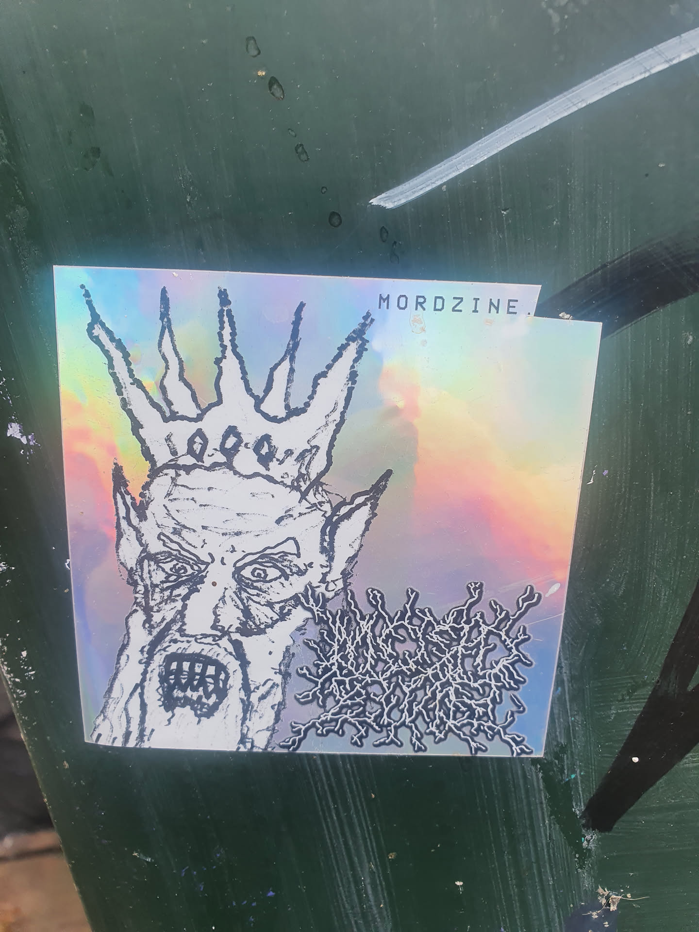 This sticker features a holographic design. The main image is a black and white drawing of a demonic or evil-looking figure wearing a crown.  There is some additional text at the bottom right, which appears to be a band logo.  The sticker is rectangular with a slight curve to the top right corner. The text 'MORDZINE.' is visible on the sticker.