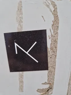A small, rectangular sticker with a dark background and a simple, white, abstract 'K' like symbol in the center. The sticker is affixed to a light gray surface that shows some wear and tear.