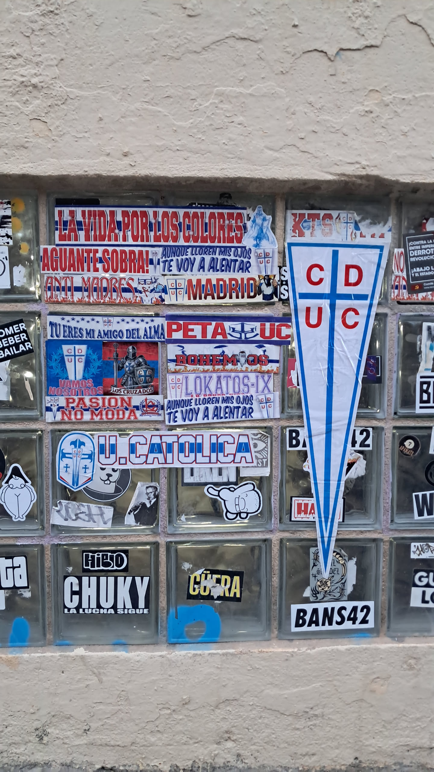 The central sticker is a large, white pennant-shaped emblem with a prominent blue border and two vertical blue stripes forming a cross. Inside the white space, the letters 'CD' are stacked vertically on the left side of the cross, and 'UC' are stacked vertically on the right side, all in bold red capital letters. The sticker shows some minor creases, particularly towards the bottom, indicating it is a physical adhesive item.