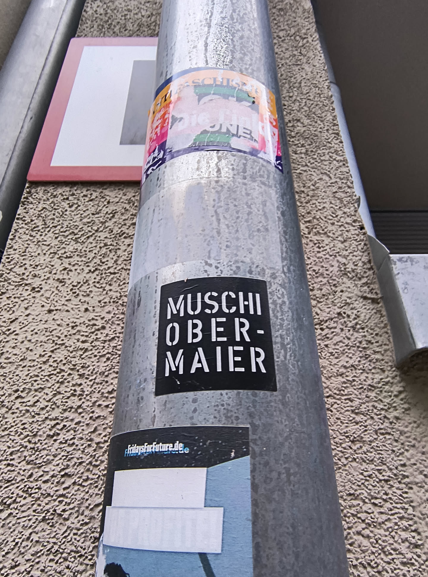 The sticker is rectangular with slightly rounded corners, featuring a solid black background. White, stencil-style uppercase letters are prominently displayed, spelling 'MUSCHI' on the top line and 'OBER-MAIER' on the bottom line. The text is centered within the sticker. The sticker shows minor signs of wear, including slight creases and some dirt, indicating its exposure to the elements on the metallic pole.