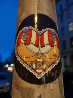 A round sticker with a heart-shaped design shows two backsides, each adorned with a red and white striped canopy.  Nautical elements, including a floral motif and a life preserver, are incorporated into the design. The style is reminiscent of vintage pin-up art or tattoo flash.