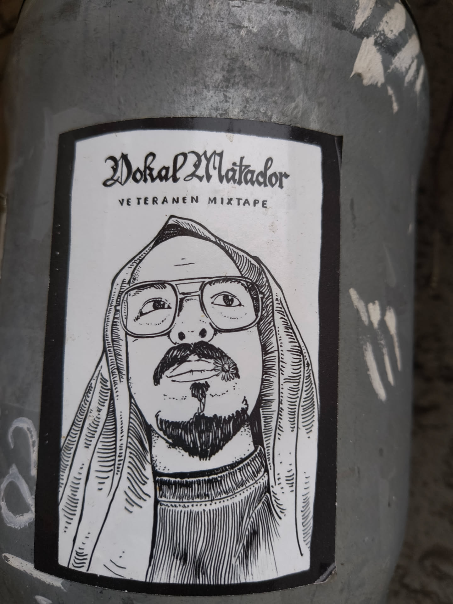 The sticker features a black and white illustration of a man's face, centered within a black border. The man is wearing glasses and has a goatee and mustache. He is also depicted wearing a hooded garment, with the hood pulled up over his head. The style of the illustration is detailed, with fine lines used to create shading and texture, particularly on the clothing and facial hair. Above the portrait, in a stylized gothic font, is the text 'Vokal Matador', and below that, in a simpler font, is 'VETERANEN MIXTAPE'.