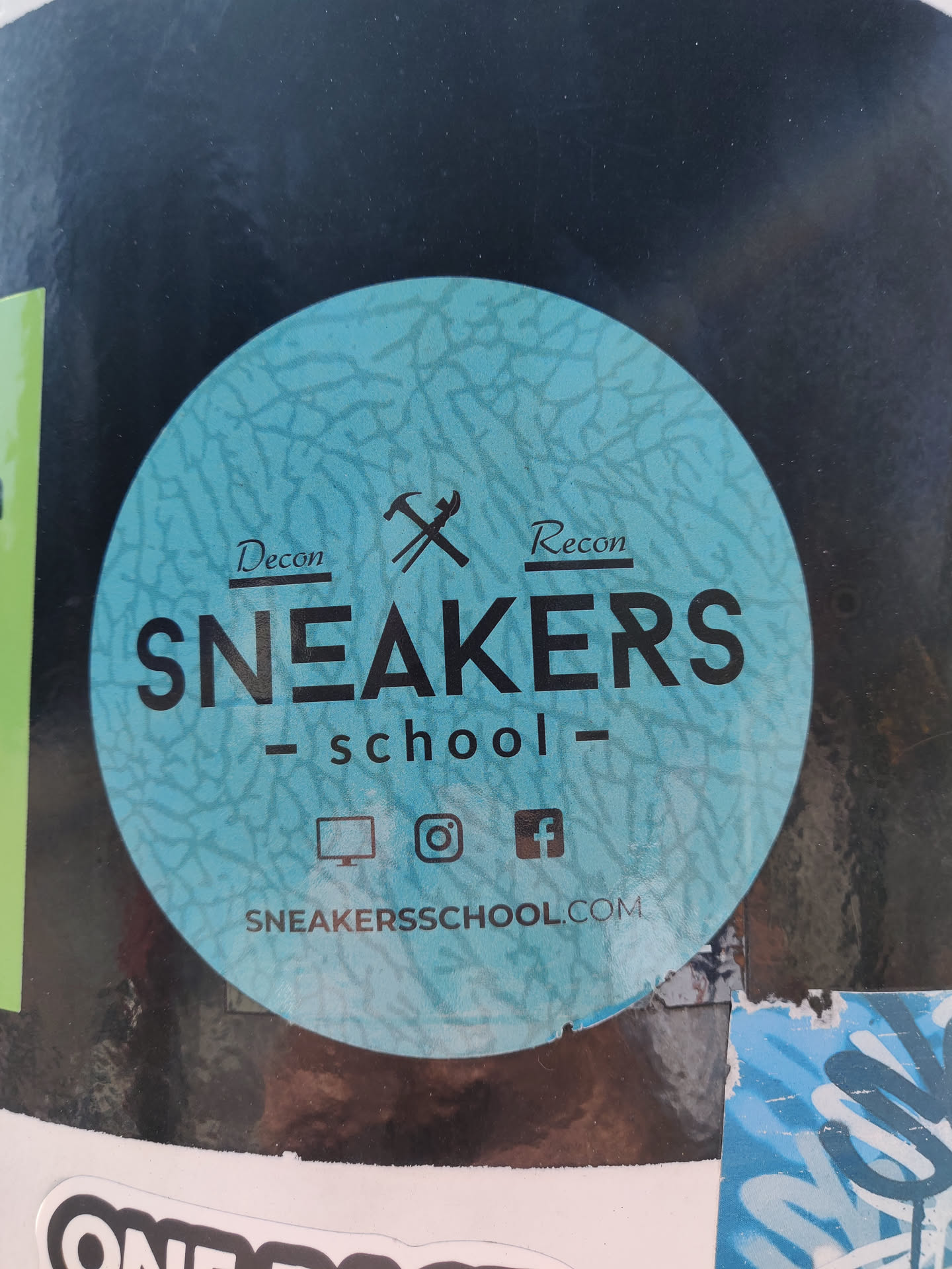 Round sticker with a teal cracked-like pattern. It has the text 'Sneakers School' in black uppercase letters with 'Decon' and 'Recon' on either side of crossed hammers. At the bottom, there are icons for a computer monitor, Instagram, and Facebook, followed by the website address 'sneakerssschool.com'.