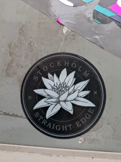 This round sticker features a pale gray lotus flower in the center, surrounded by the text "STOCKHOLM STRAIGHT EDGE" in a circular pattern. The background is a darker gray, and small plus signs are placed on either side of the word "STOCKHOLM".