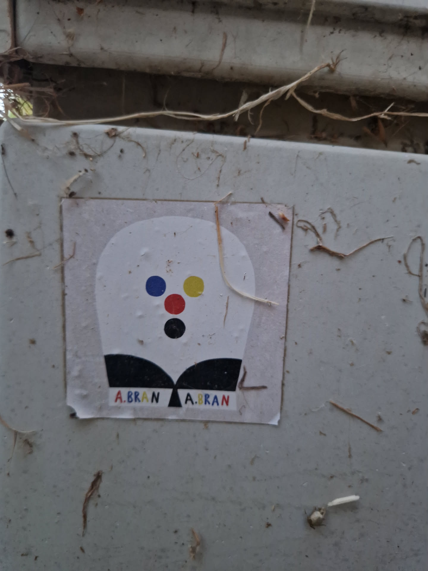 A small, rectangular sticker with a simple design. The main element is a stylized head-like shape with a few colorful dots and a black bottom. The text 'A.BRAN' is repeated twice at the bottom of the sticker.