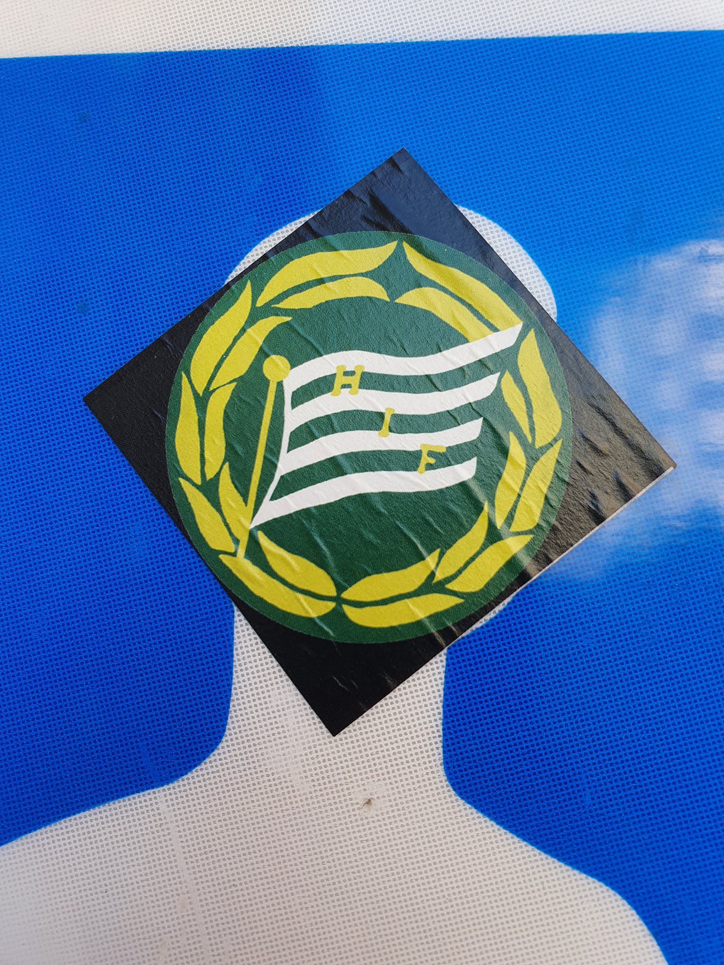 A square sticker featuring a green and white flag with the letters HIF in the center, surrounded by a yellow laurel wreath. The sticker is adhered to a blue surface. The flag appears to be slightly damaged.