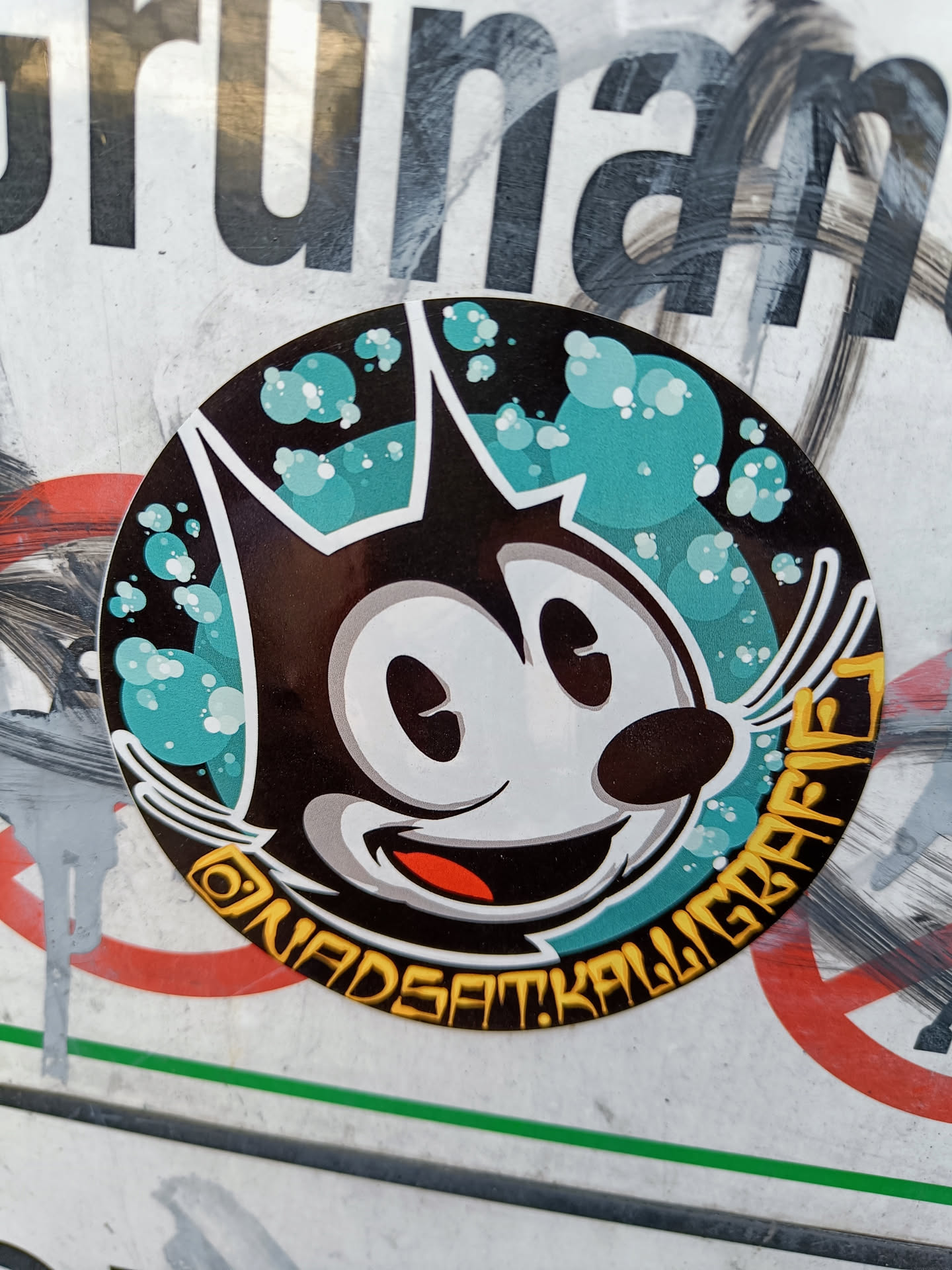 Round sticker featuring a retro-style cartoon cat with teal and white accents. The text around the cat says "ONADSATKALIGRAFI".