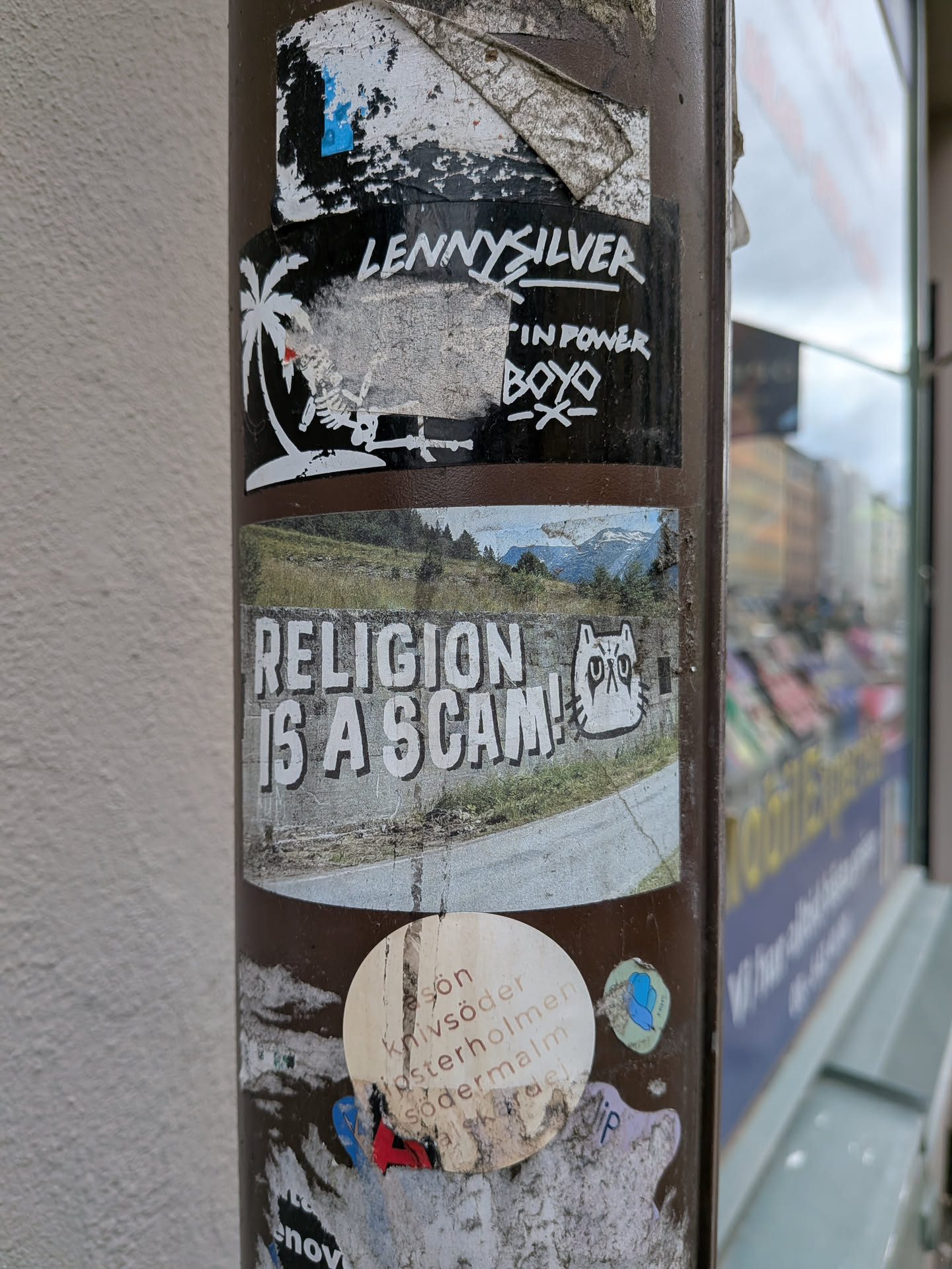 A rectangular sticker featuring a composite image. The background is a scenic landscape with a winding road in the foreground, green hills and trees, and snow-capped mountains under a cloudy sky in the distance. Overlaid on this landscape, in large, bold white letters with a dark drop shadow, is the phrase 'RELIGION IS A SCAM!'. To the right of the text, a stylized white cat head with dark, somewhat grumpy-looking eyes and mouth is depicted, with the letters 'DXO' beneath it. The sticker shows signs of wear and tear, particularly around its edges.