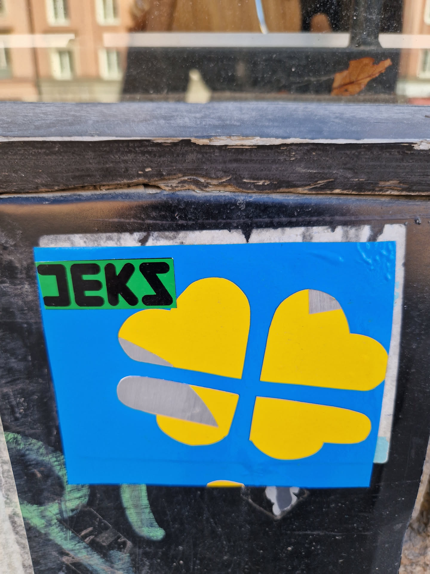 Close-up of a square sticker affixed to a dark surface. The sticker is predominantly blue, featuring a stylized yellow four-leaf clover.  A smaller green rectangle with the word "JEKS" written in black letters is visible on the sticker. 