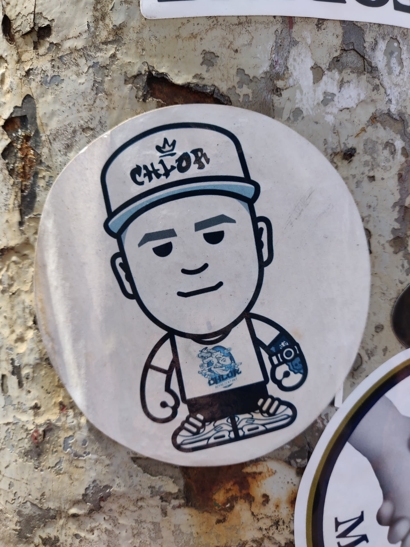 Round sticker featuring a cartoon character wearing a hat with 'CHLOF' written on it. The character has a simple design and is wearing a t-shirt with a small logo. The sticker has a slightly worn look, suggesting it has been placed outdoors for a while. The background is a weathered surface, possibly a wall or pole.