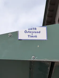 A rectangular white sticker with a blue border, attached to a green metal surface. The sticker displays the names "Östersund" and "Timrå" in blue text, possibly indicating support for these locations or the teams representing these locations.