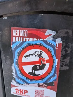 The sticker is circular with a red and white prohibition symbol superimposed on a background of blue and white wavy lines. Encircling the prohibition symbol is a jagged line resembling barbed wire. Inside the prohibition circle, a black fist is shown crushing a weapon that appears to be a rifle or gun with a bayonet. The fist itself is white and outlined in black. Below the prohibition symbol, a black silhouette of a helmet or cap with a small emblem of a dome and spire is visible. The sticker is partially overlapped by another poster with red and white text, and a QR code and text "RKP REVOLUTIONÄRA KOMMUNISTISKA PARTIET" are visible below the central sticker.
