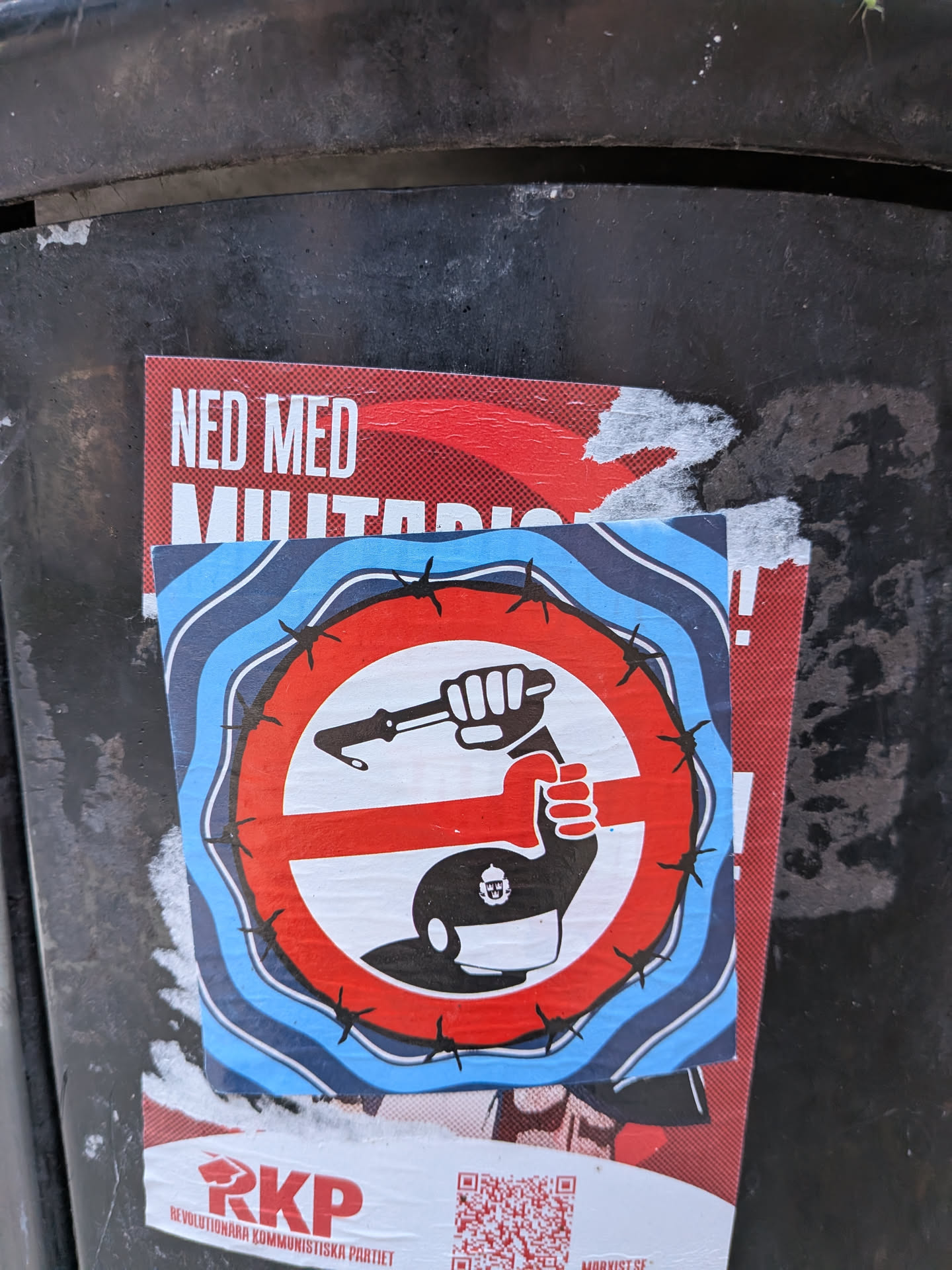 The sticker is circular with a red and white prohibition symbol superimposed on a background of blue and white wavy lines. Encircling the prohibition symbol is a jagged line resembling barbed wire. Inside the prohibition circle, a black fist is shown crushing a weapon that appears to be a rifle or gun with a bayonet. The fist itself is white and outlined in black. Below the prohibition symbol, a black silhouette of a helmet or cap with a small emblem of a dome and spire is visible. The sticker is partially overlapped by another poster with red and white text, and a QR code and text "RKP REVOLUTIONÄRA KOMMUNISTISKA PARTIET" are visible below the central sticker.