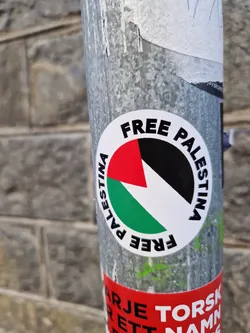 A round sticker with the text "FREE PALESTINA" written around a stylized Palestinian flag. The sticker is attached to a metal pole.