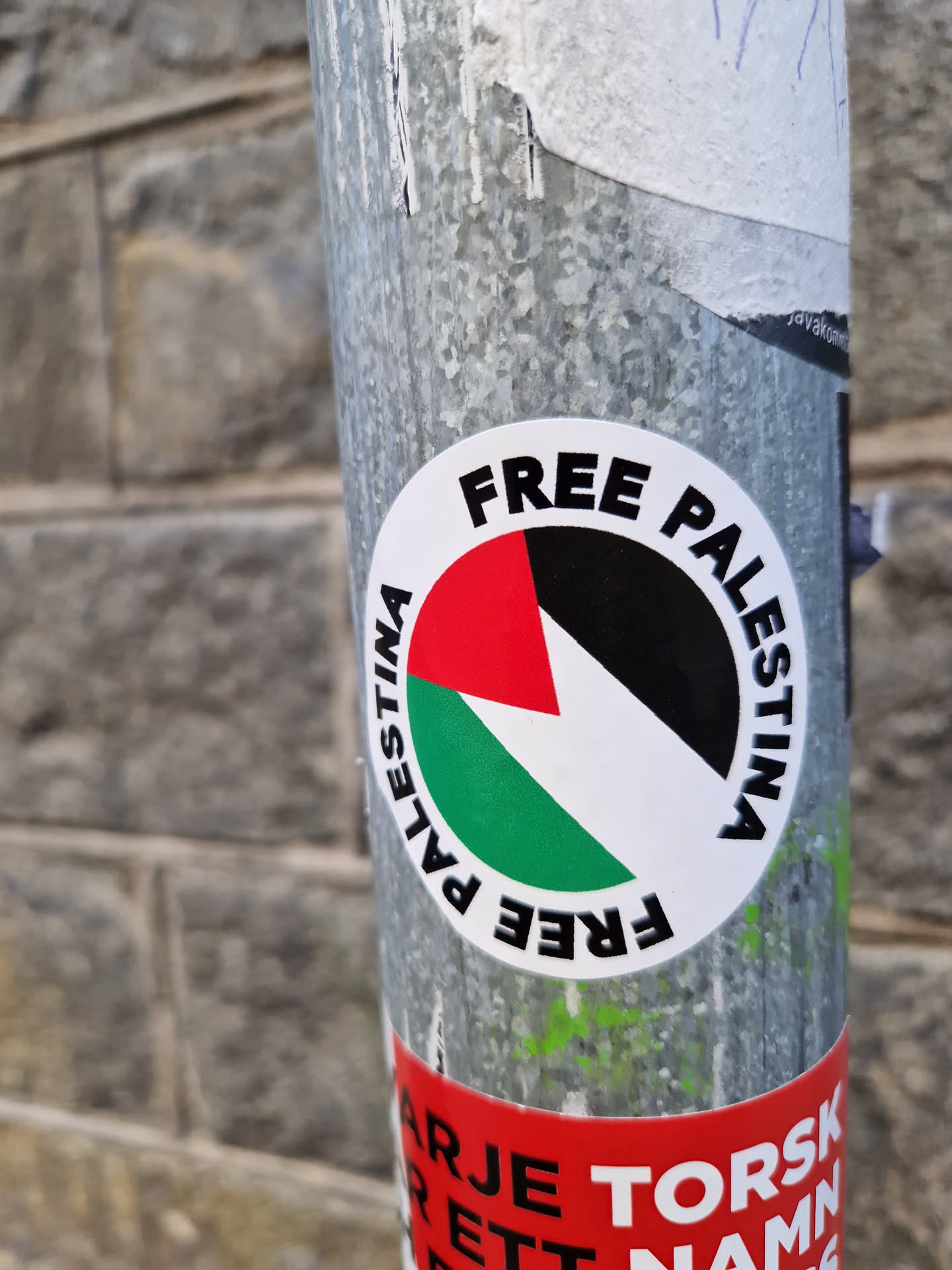 A round sticker with the text "FREE PALESTINA" written around a stylized Palestinian flag. The sticker is attached to a metal pole.