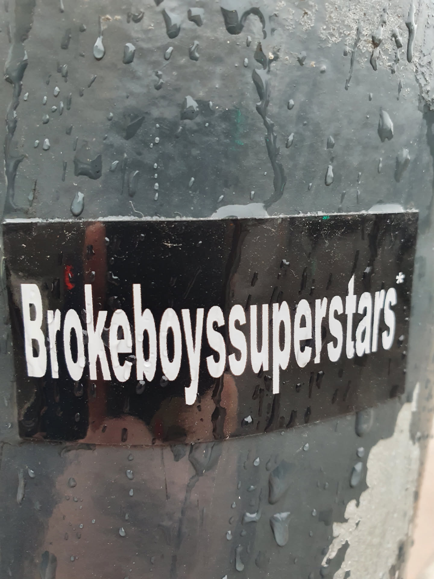 A black sticker with the text "Brokeboysuperstars" in white, slightly reflective. It's affixed to a dark-colored, rain-wet surface.