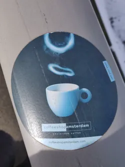 Round sticker with a dark blue background featuring a light blue espresso cup with steam rings above it. The text "coffeeshopamsterdam" is prominently displayed at the bottom, along with the tagline "amsterdam coffee" and website address "coffeeshopamsterdam.com".