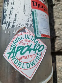 A diamond-shaped sticker with the text "APO440" in a stylized graffiti font. The words "TRAVEL ULTRAS", "BULL TOWN CITY", "STREETART", and "WORLDWIDE" surround the central text. The sticker is white with red and green accents, and it's attached to a metal pole.