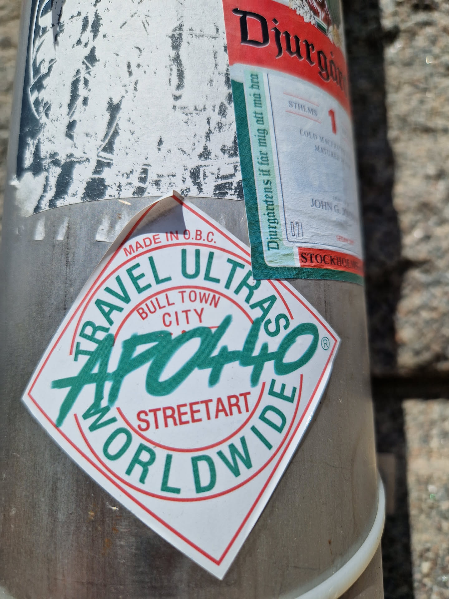 A diamond-shaped sticker with the text "APO440" in a stylized graffiti font. The words "TRAVEL ULTRAS", "BULL TOWN CITY", "STREETART", and "WORLDWIDE" surround the central text. The sticker is white with red and green accents, and it's attached to a metal pole.