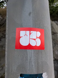 The sticker is a horizontally oriented rectangle with a vibrant red background. Centered on this background are large, interconnected white shapes that form a highly stylized, abstract symbol. The shapes are thick and rounded, creating a cohesive design that could be interpreted as a monogram or a unique emblem. The sticker is affixed to a textured grey metal pole and shows visible water droplets on its surface, indicating outdoor exposure.