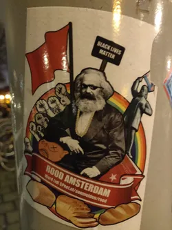 This sticker features Karl Marx in the center, with raised fists, a red flag, a Black Lives Matter sign, and a rainbow in the background.  There are loaves of bread at the bottom.  The text at the bottom says "ROOD AMSTERDAM" and includes a URL.