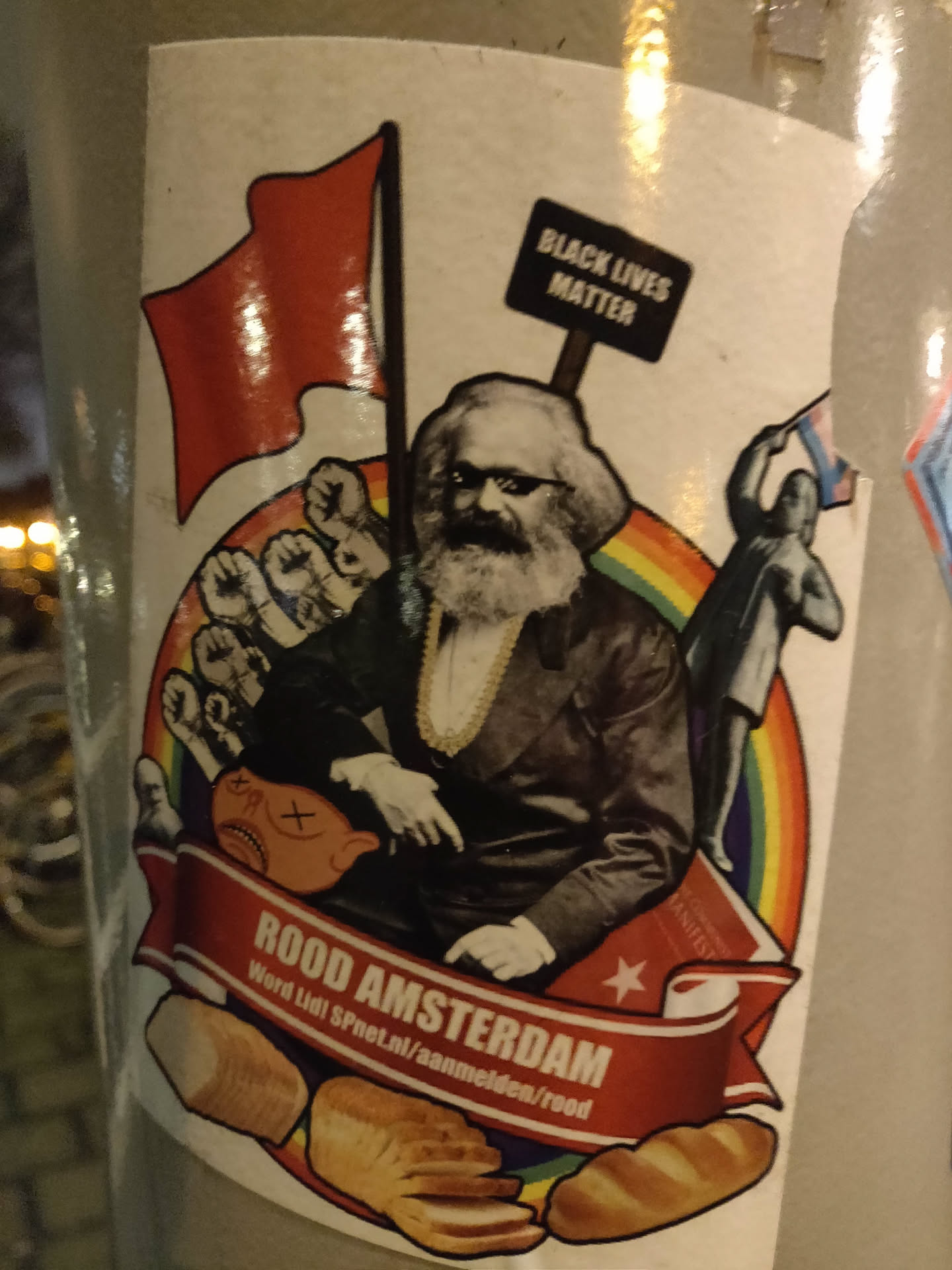 This sticker features Karl Marx in the center, with raised fists, a red flag, a Black Lives Matter sign, and a rainbow in the background.  There are loaves of bread at the bottom.  The text at the bottom says "ROOD AMSTERDAM" and includes a URL.