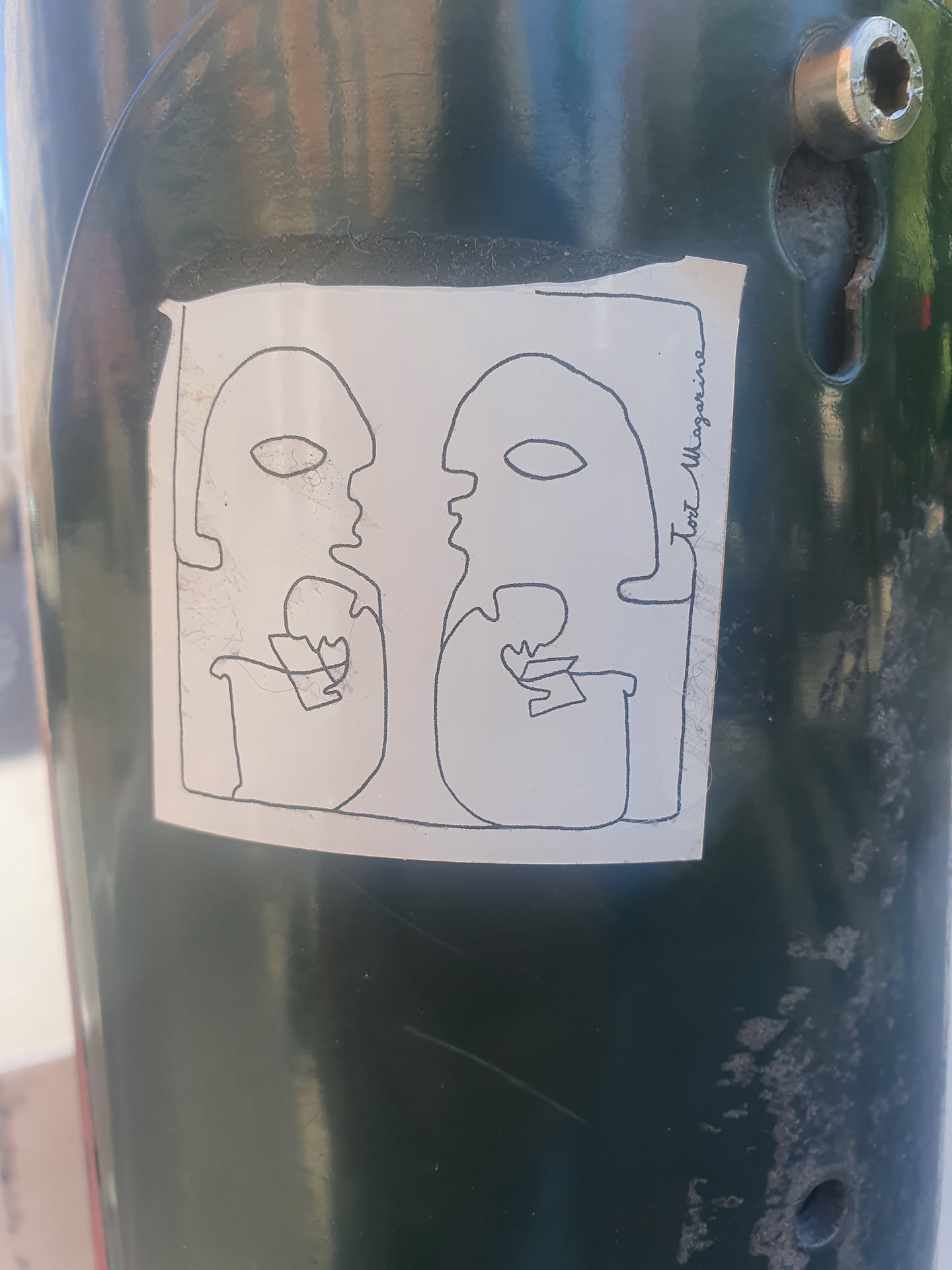 A minimalist line drawing sticker of two faces seemingly in conversation, with the text 'Cool Magazine' subtly placed on the side. The sticker is attached to a dark green pole.